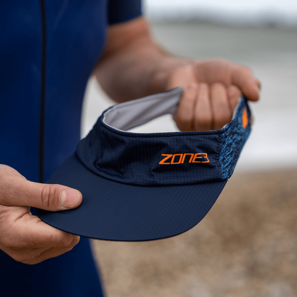 Navy lightweight race visor with orange Zone3 logo held by a swimmer on a beach.