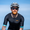 Man cycling outdoors wearing the ZONE3 Activate+ men’s short sleeve trisuit in black, with blue sleeve cuffs and an orange chest logo.
