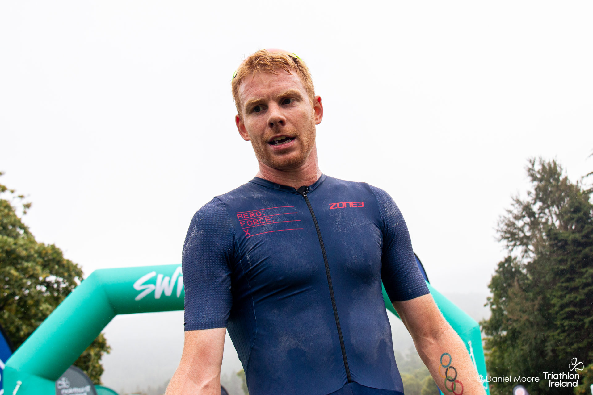 Russell White in a navy ZONE3 trisuit at a race, with an event arch behind.