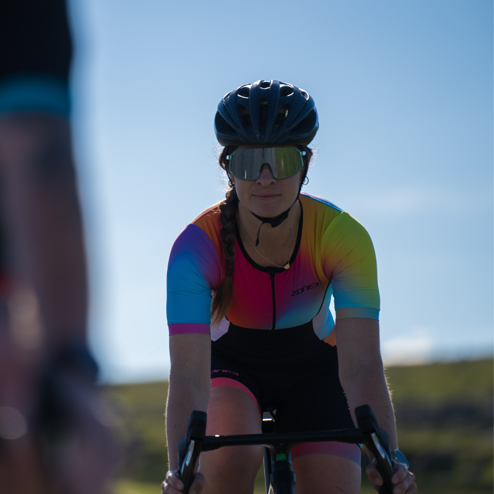 Woman cycling head-on wearing the ZONE3 Activate+ women’s short sleeve trisuit with mirrored sunglasses and a black helmet. Gradient design clearly shown across the chest and sleeves.