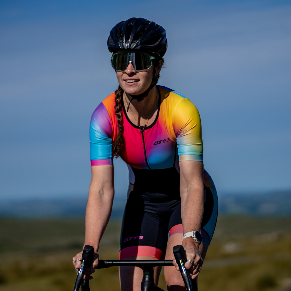 Close-up front view of a cyclist wearing the ZONE3 Activate+ women's short sleeve trisuit. Gradient colours in blue, pink, yellow, and orange are visible under sunlight.