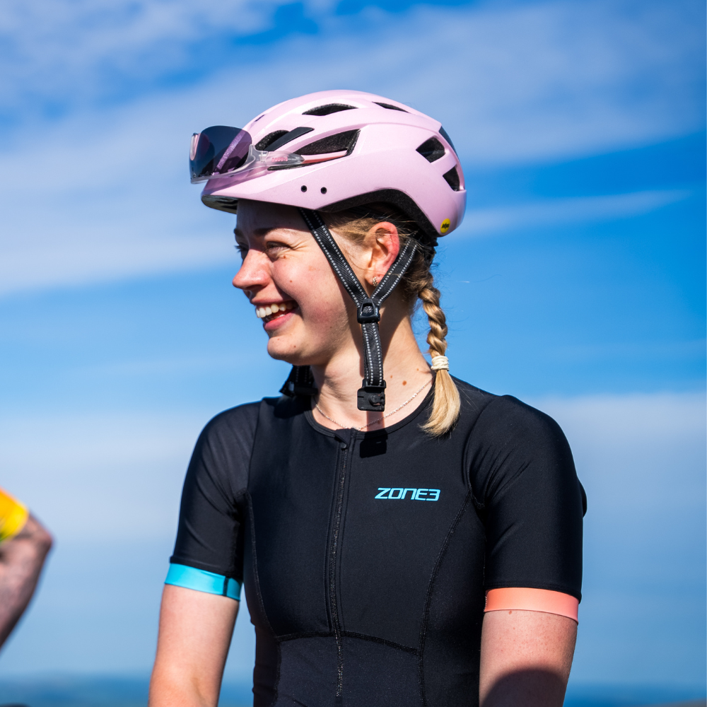 Woman wearing the ZONE3 Activate+ women’s short sleeve trisuit in black with pink and blue sleeve cuffs. She is smiling and wearing a pink helmet.
