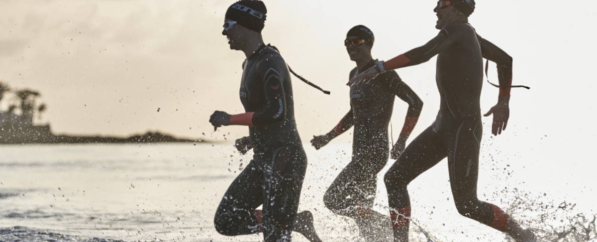Three swimmers in wetsuits running into the sea together.