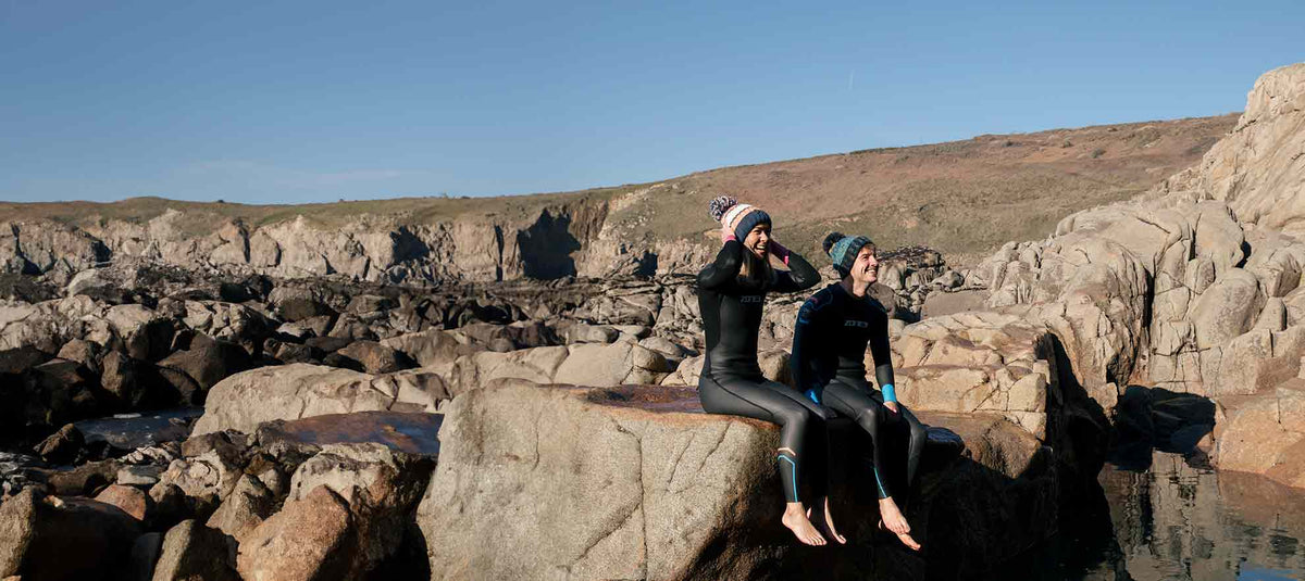 Two swimmers in wetsuits sitting together on rocks above the sea.