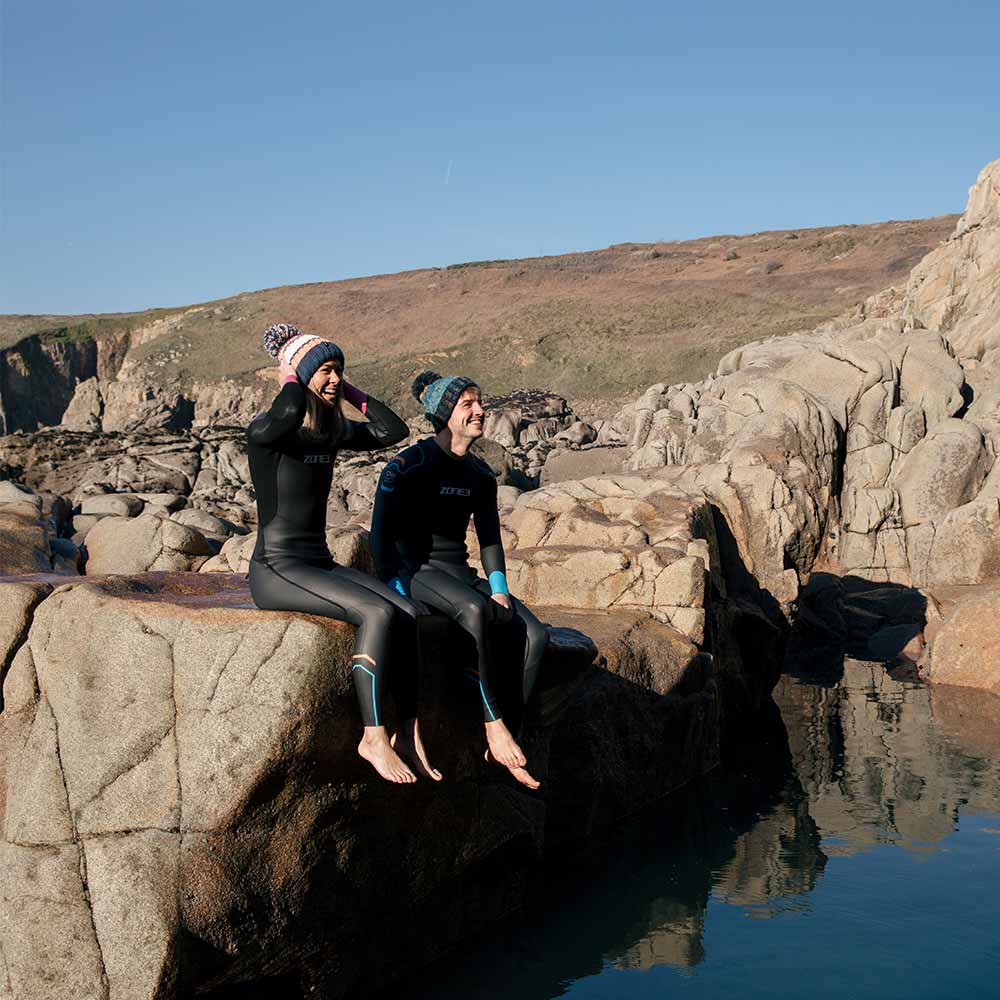 Two swimmers in wetsuits sitting together on rocks above the sea.