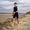 Person wearing blue Yulex kneeskin wetsuit walks on a beach holding an orange water bottle